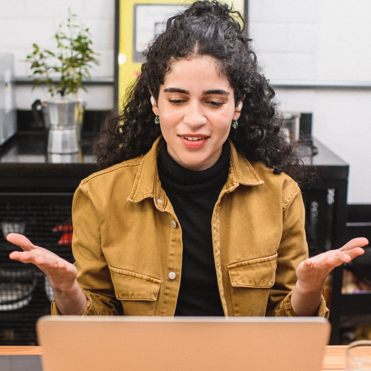 Uma mulher gesticula com as mãos aberta em frente a um computador enquanto sorri. O fundo da foto é uma cozinha desfocada.